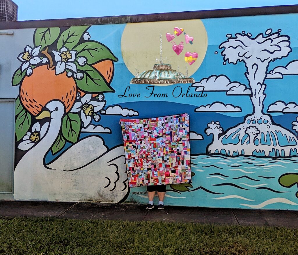 Me holding up the finished crumb quilt in front of an Orlando mural that shows oranges, a swan and the Lake Eola fountain.