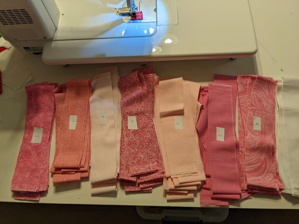 2.5" strips of varying hues and designs of pink fabric laid out in front of my sewing machine.