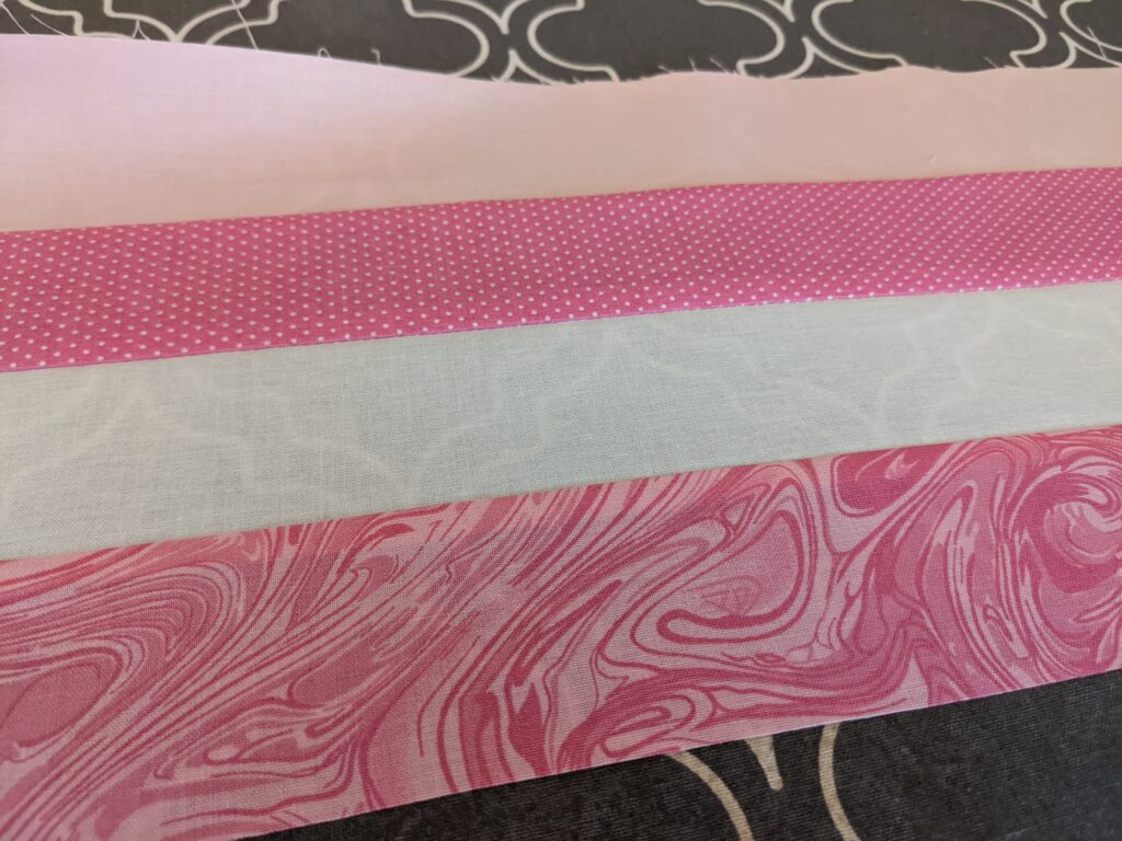 Sewing strip set with 4 different fabrics in hues of pink laid out on top of my ironing board.