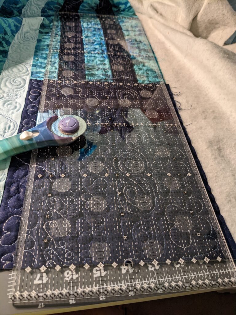 Quilting ruler on top of the quilt with a rotary cutter on top of both.