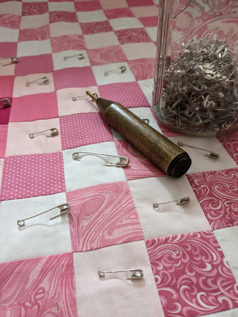 Close up of my Kwik Klip tool, with a jar of safety pins, on top of the Around the World quilt top that is partially basted with safety pins.