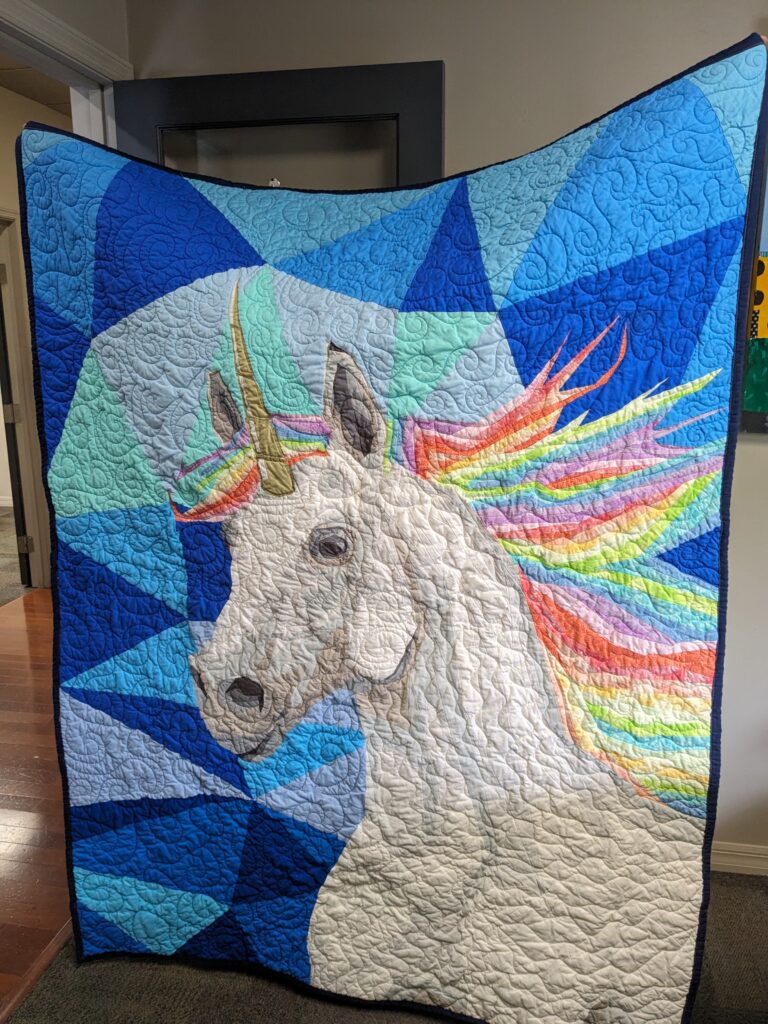 Unique-orn quilt that I made. It is a unicorn with a rainbow mane with varying blue fabrics for the background. It is quilted with mainly swirls.